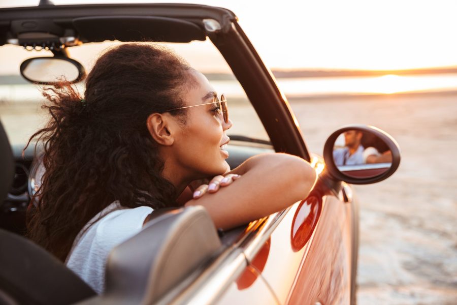 Image of young pretty woman smiling and looking aside while riding in convertible stylish car by seaside
